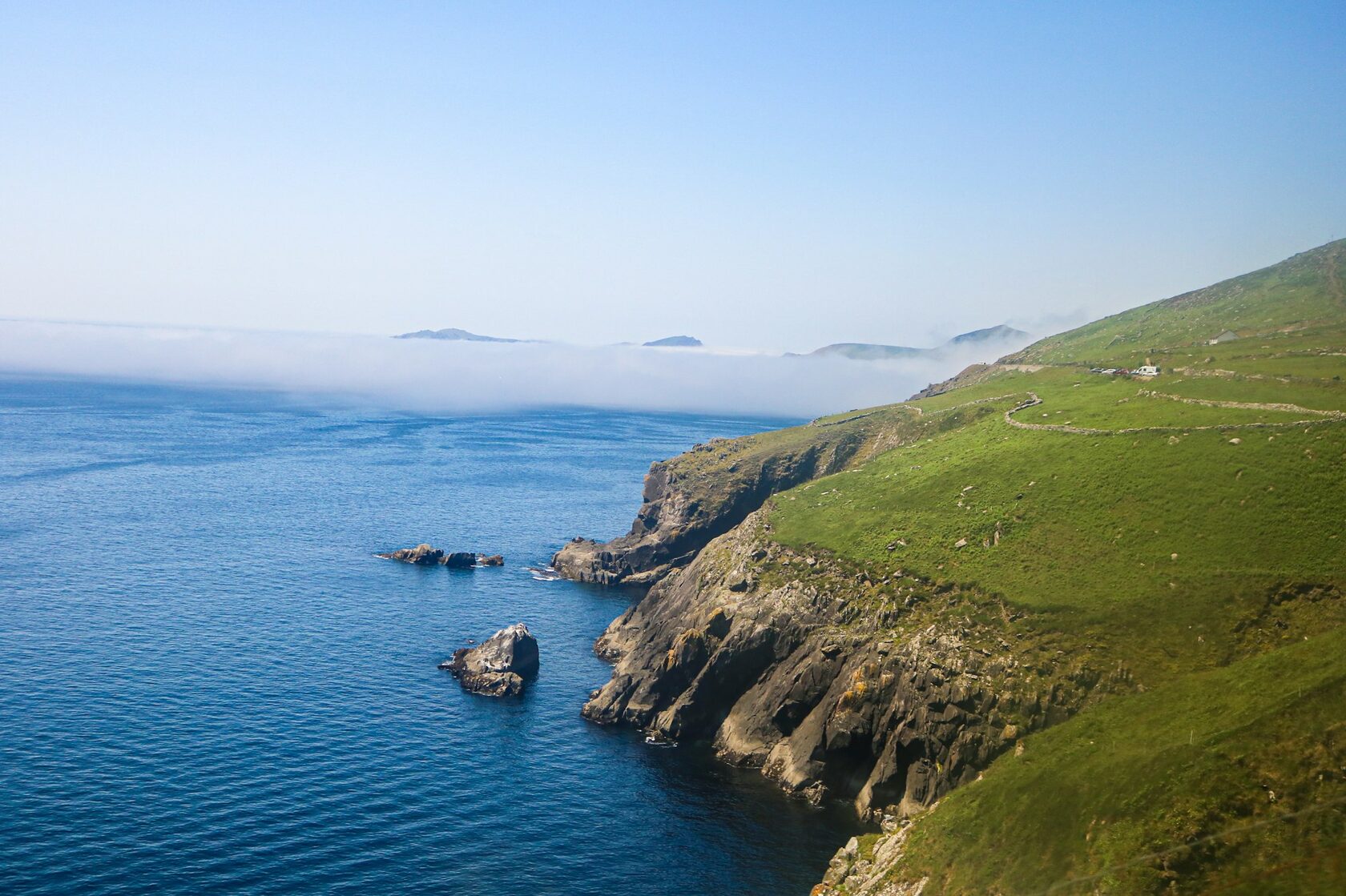 Dingle Peninsula, Ireland, at Slea Head in golden hour light. Wide view of green fields and old stone walls rolling down to the Atlantic, with the Blasket Islands on the horizon.