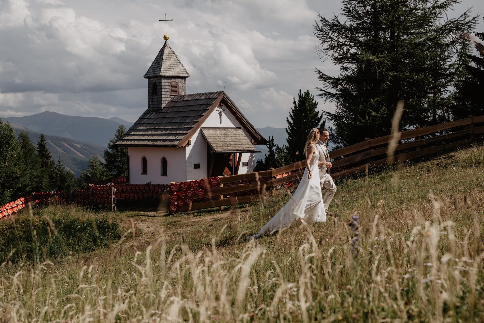 Bride in a flowing whote wedding dress walking through a sunlit alpine meadow beside a small mountain chapel in Obertauern