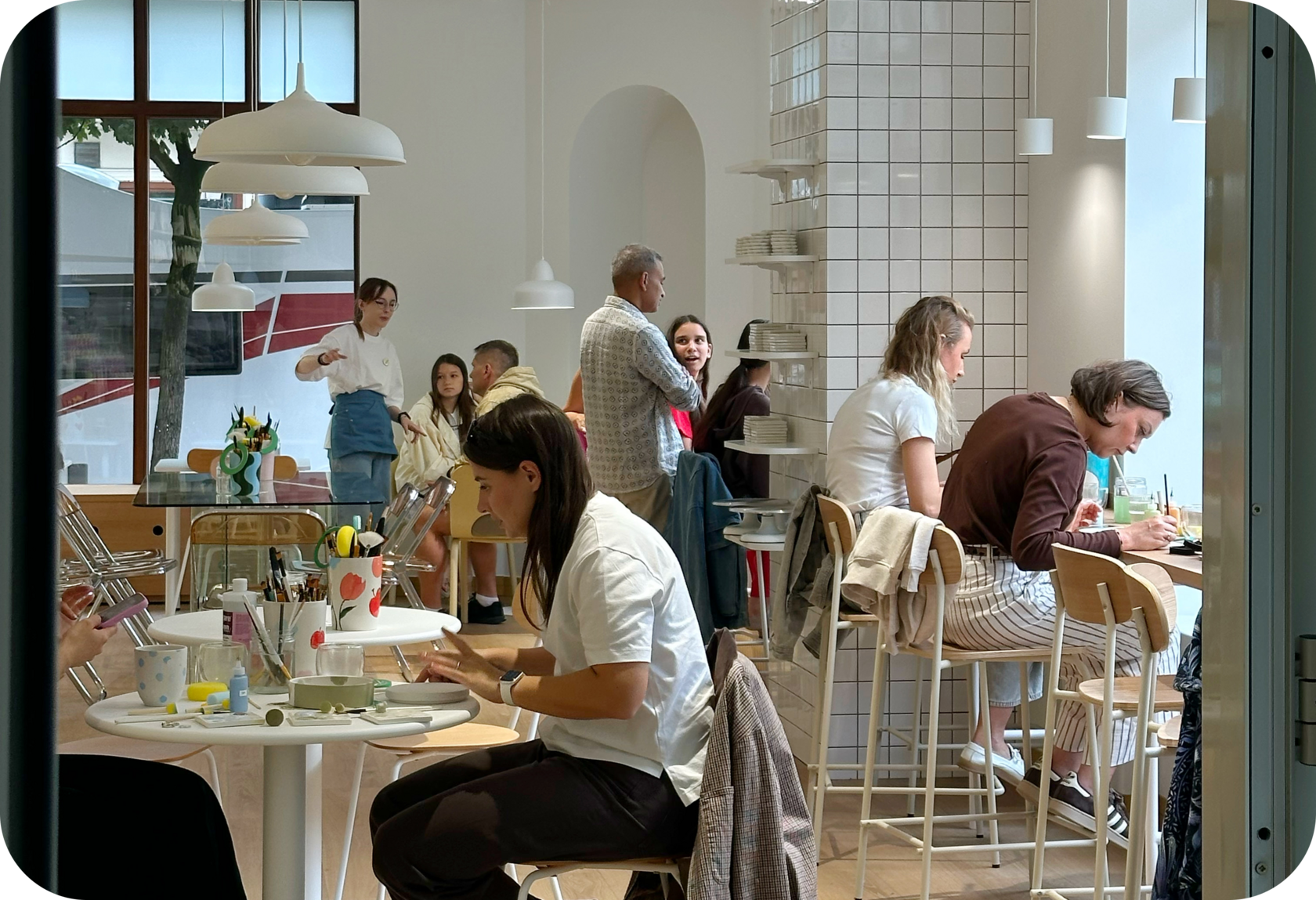 Colleagues painting ceramics at round tables in Create Cafe Poznań — a bright interior with a tiled column and an arched niche