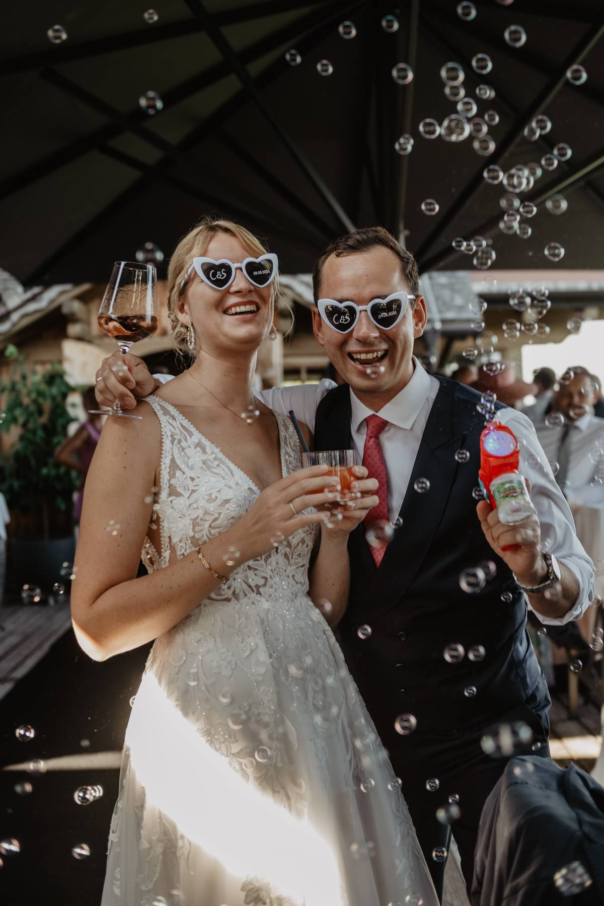 Bride and groom laughing in heart-shaped sunglasses at a mountain wedding reception, bubbles and drinks in hand