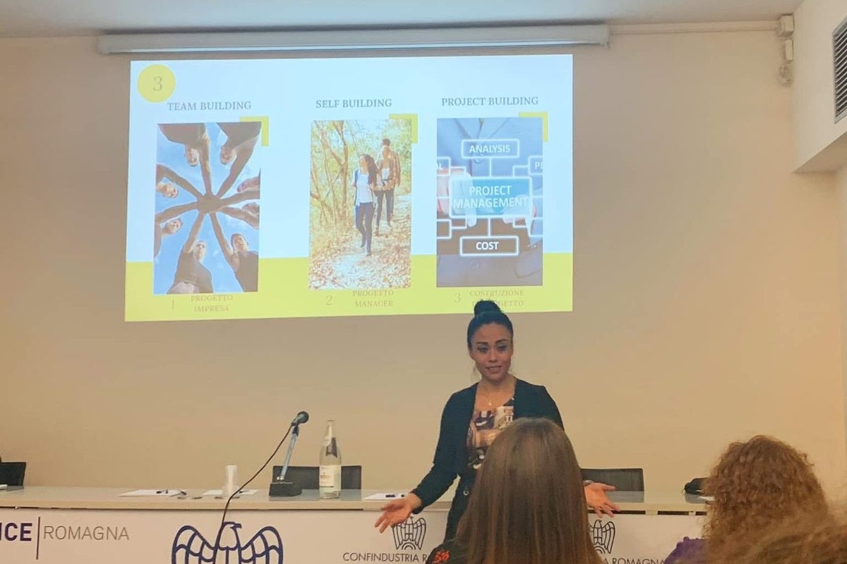 Ana Laura Ramirez in piedi davanti allo schermo mentre parla a una sala, al tavolo dei relatori il marchio di Confindustria Romagna