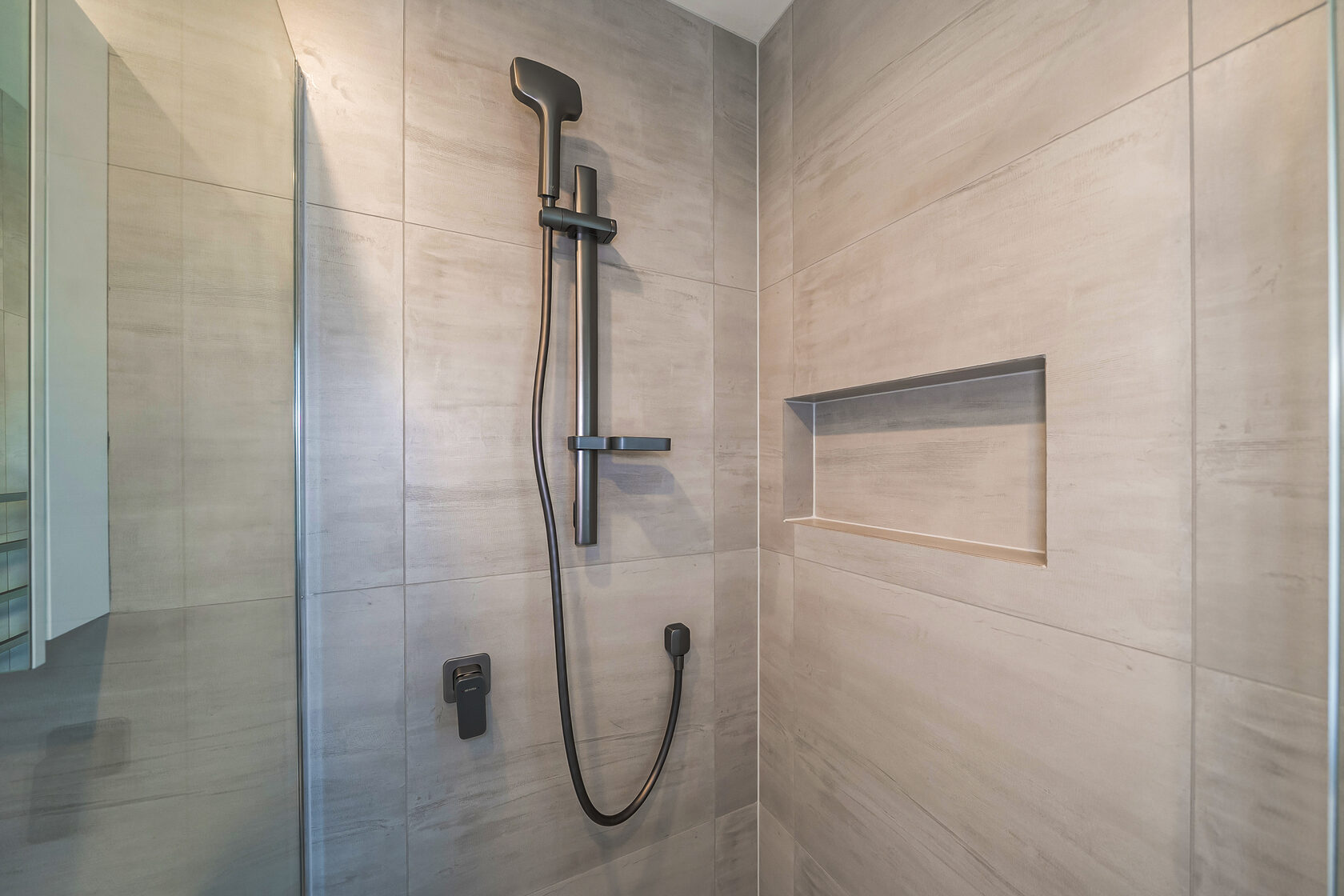 Modern walk-in shower featuring large-format beige tiles, matte black shower rail and handheld shower head, built-in wall niche for storage, and frameless glass panel.