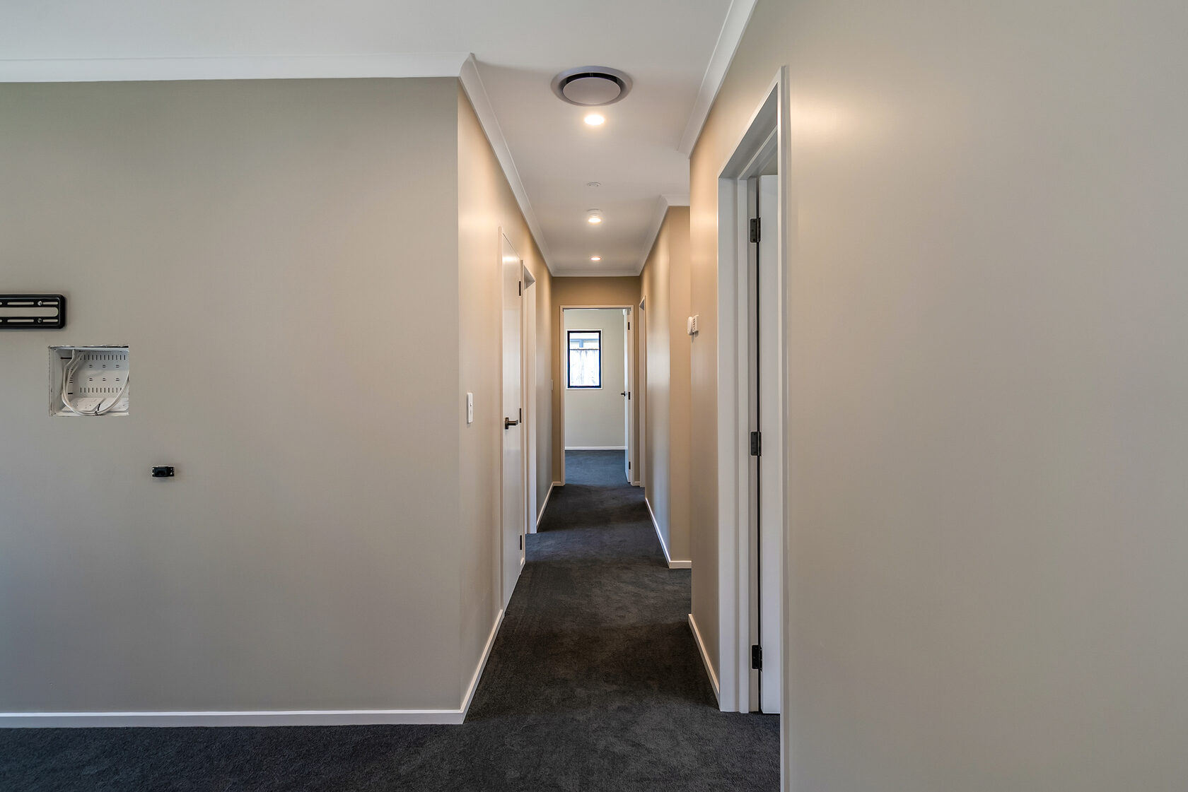 Long interior hallway with neutral walls, dark carpet, recessed ceiling lights, and multiple doorways leading to adjoining rooms.