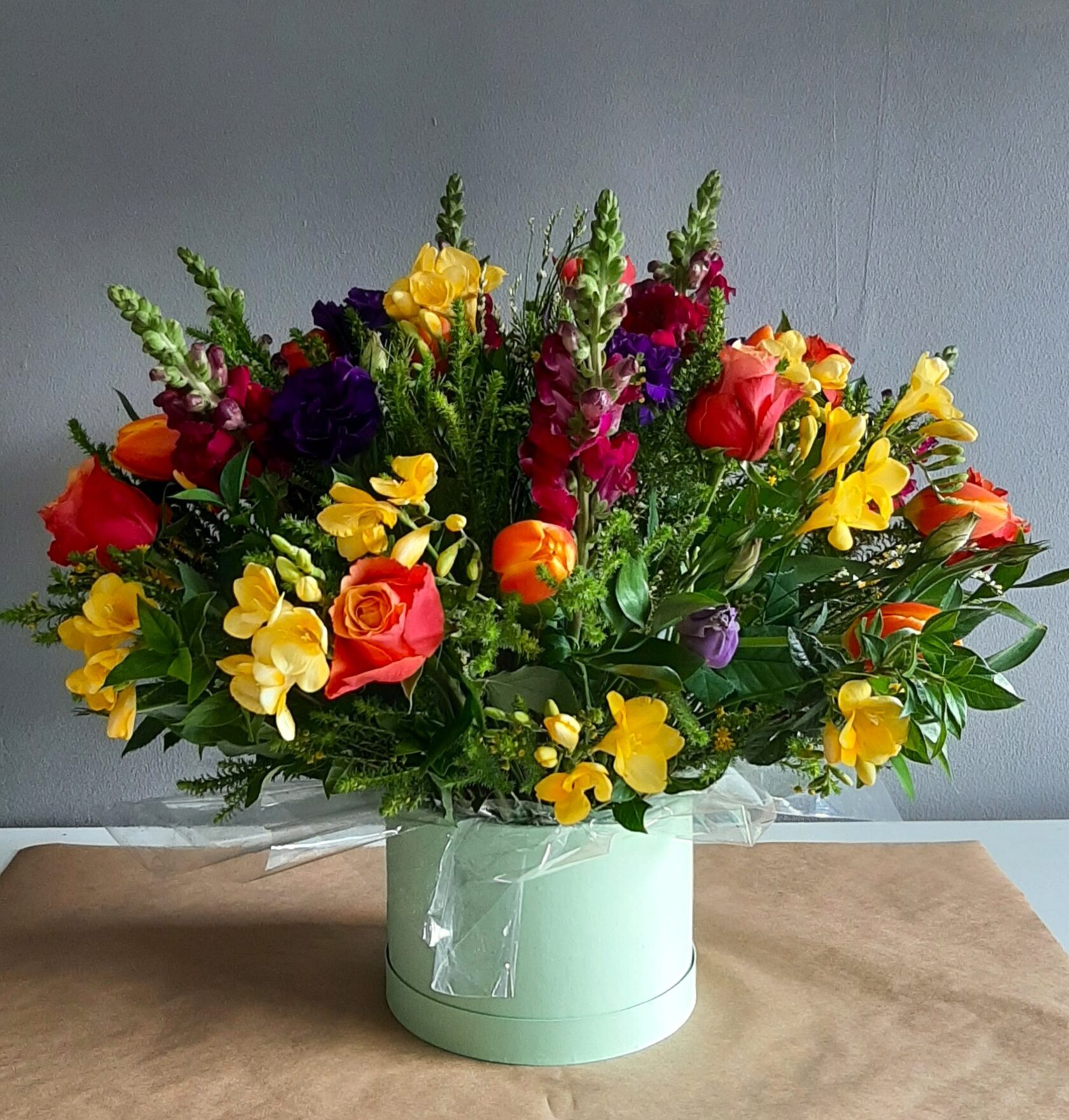 A vibrant mixed floral arrangement in a light green hat box, featuring roses, snapdragons (antirrhinum), freesias, and other seasonal flowers.