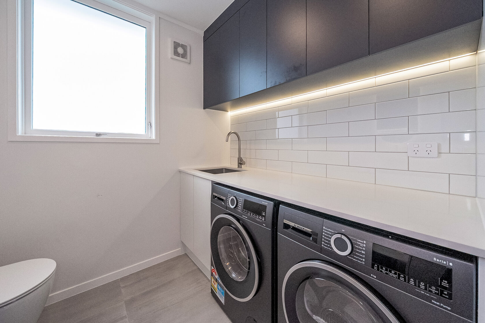 Modern laundry room with double front-loading washer and dryer, white subway tile splashback, and overhead cabinetry with LED lighting.