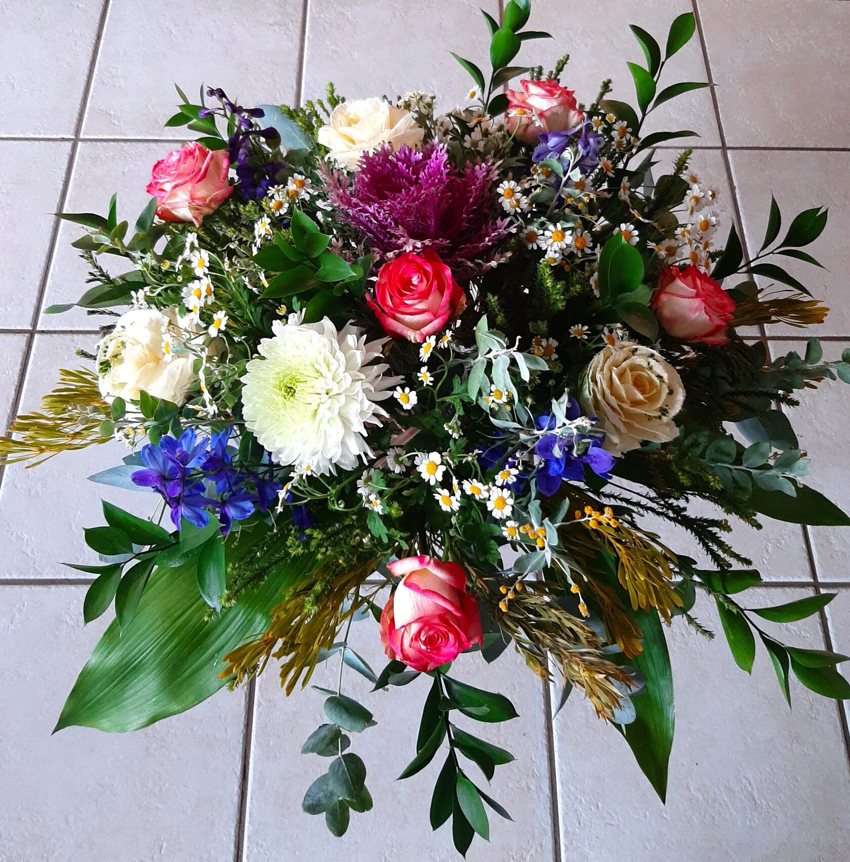 A lush bouquet featuring bright pink roses, white chrysanthemums, and field daisies, complemented by blue flowers and assorted greenery.
