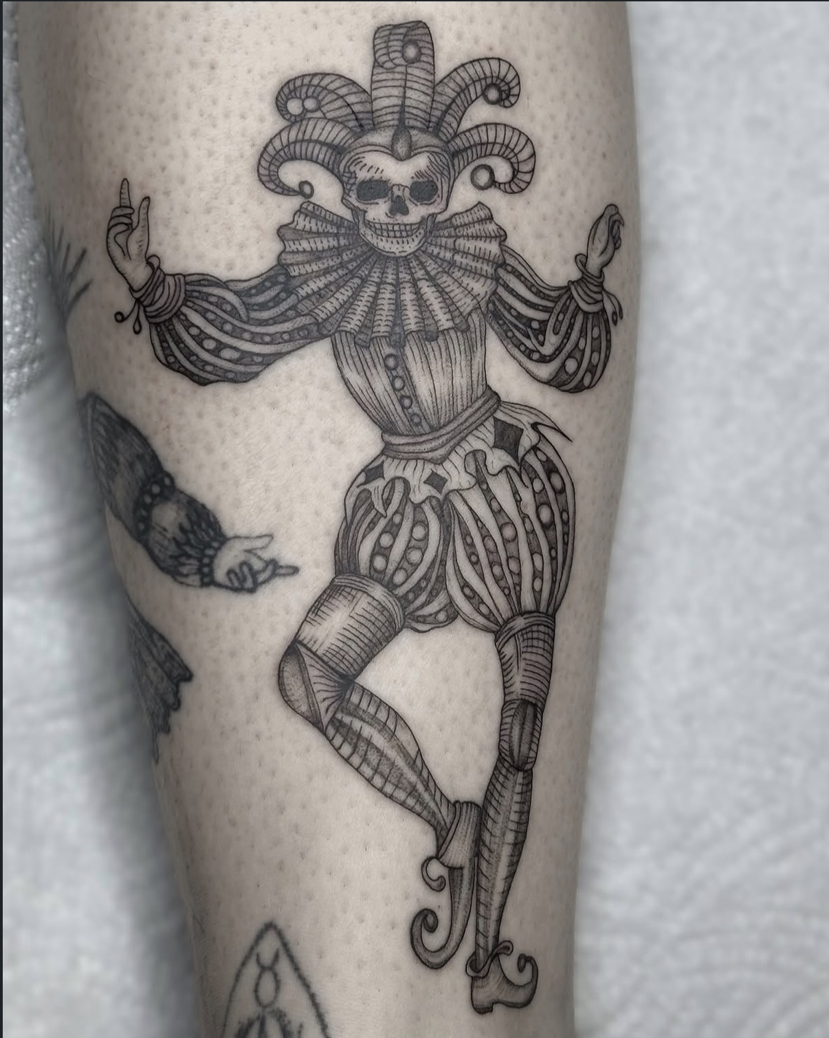 Leg tattoo of a jester with a skull face, multi-point cap, ruff collar, striped costume, raised hands and pointed shoes.