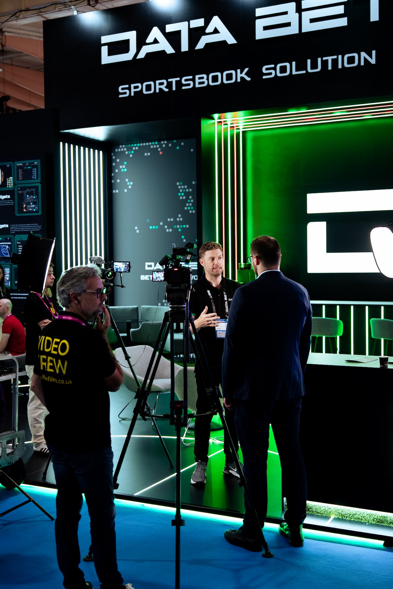 A crew filming an interview at a trade show stand, camera on a tripod beside a softbox