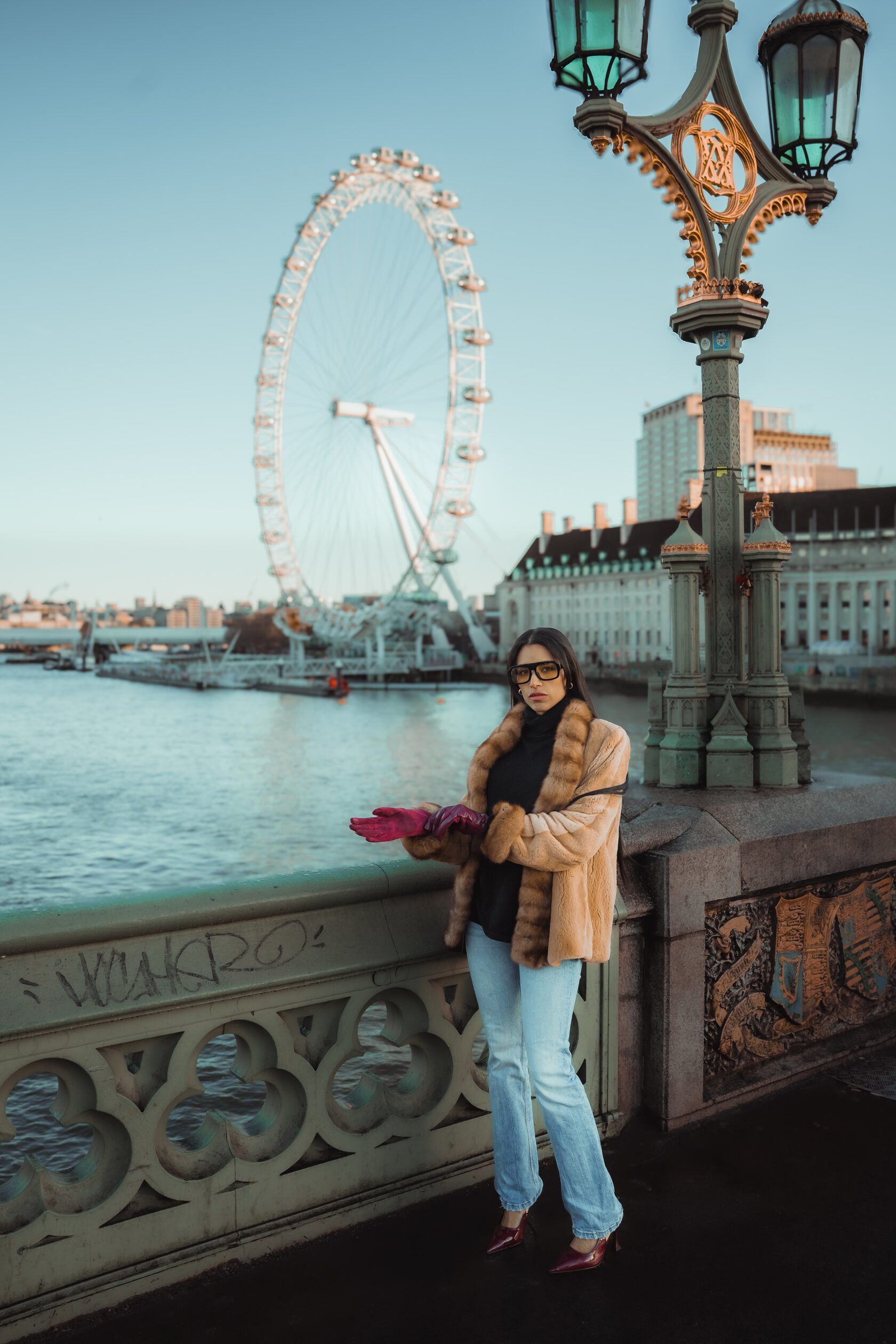 Woman in a camel jacket beside Westminster Bridge with the London Eye behind