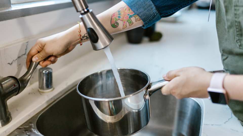 The image shows a person filling a pot with tap water in a kitchen, highlighting concerns about water quality in Cyprus and the importance of proper filtration