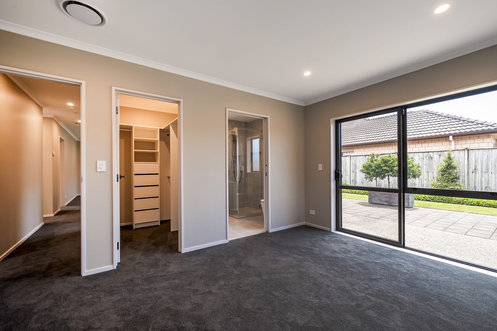 Bright master bedroom with dark carpet and neutral walls, featuring a walk-in wardrobe on the left, a private ensuite bathroom straight ahead, and large sliding glass doors opening to a patio and fenced backyard.