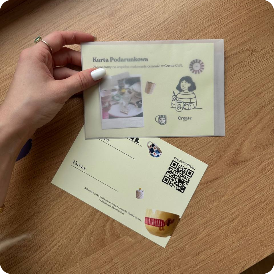 Wydrukowana karta podarunkowa Create Cafe z kodem QR w dłoni, na drewnianym stole