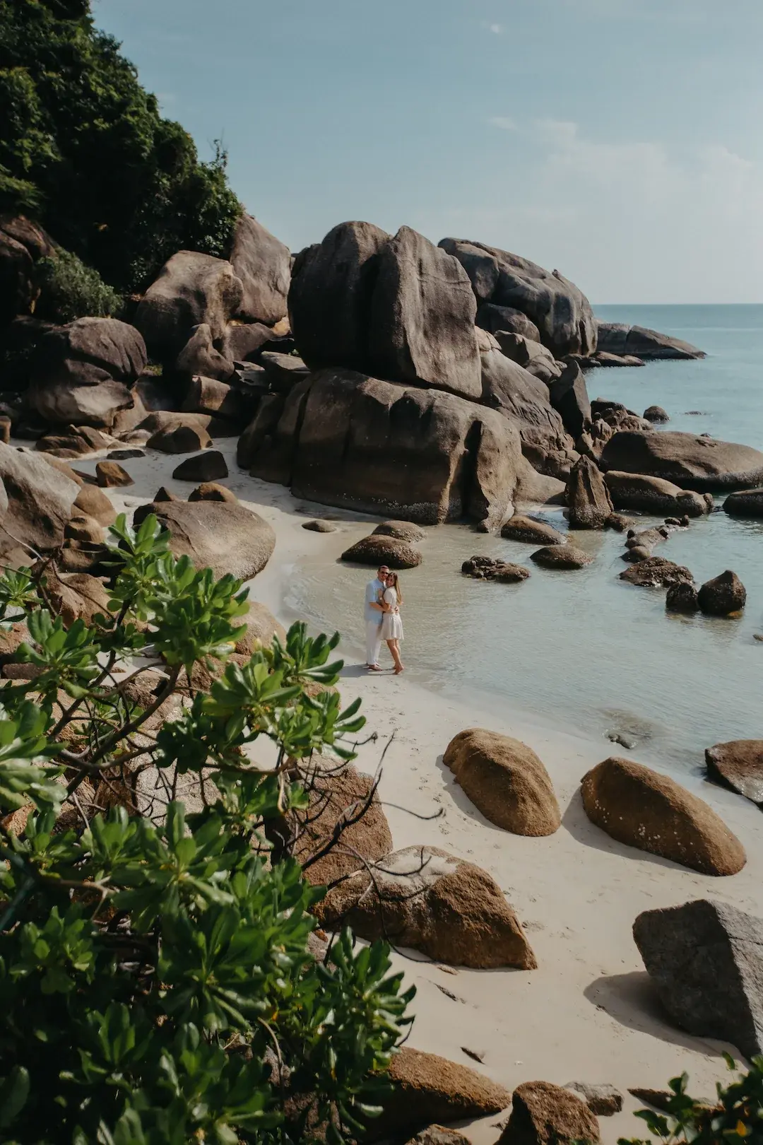 Romantic couple standing together on a secluded sandy beach with large granite boulders and calm sea at Silver Beach in Koh Samui