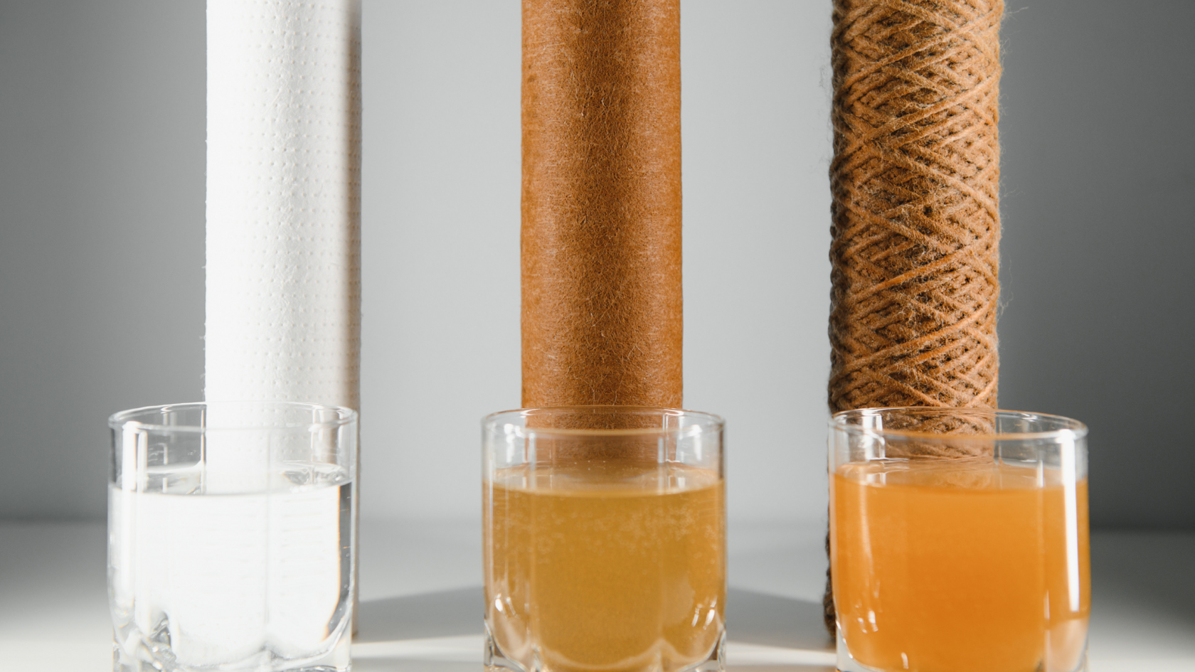 The image shows a comparison of water filtration results: three different water filter cartridges with three glasses of water, each displaying a different level of contamination. The left cartridge is clean, with a glass of clear water next to it. The middle cartridge is partially used, and the water in the glass has a slightly yellowish tint. The right cartridge is heavily contaminated, and the water in the glass has a dark orange, murky appearance. This illustrates the effectiveness and potential drawbacks of reverse osmosis systems for home use