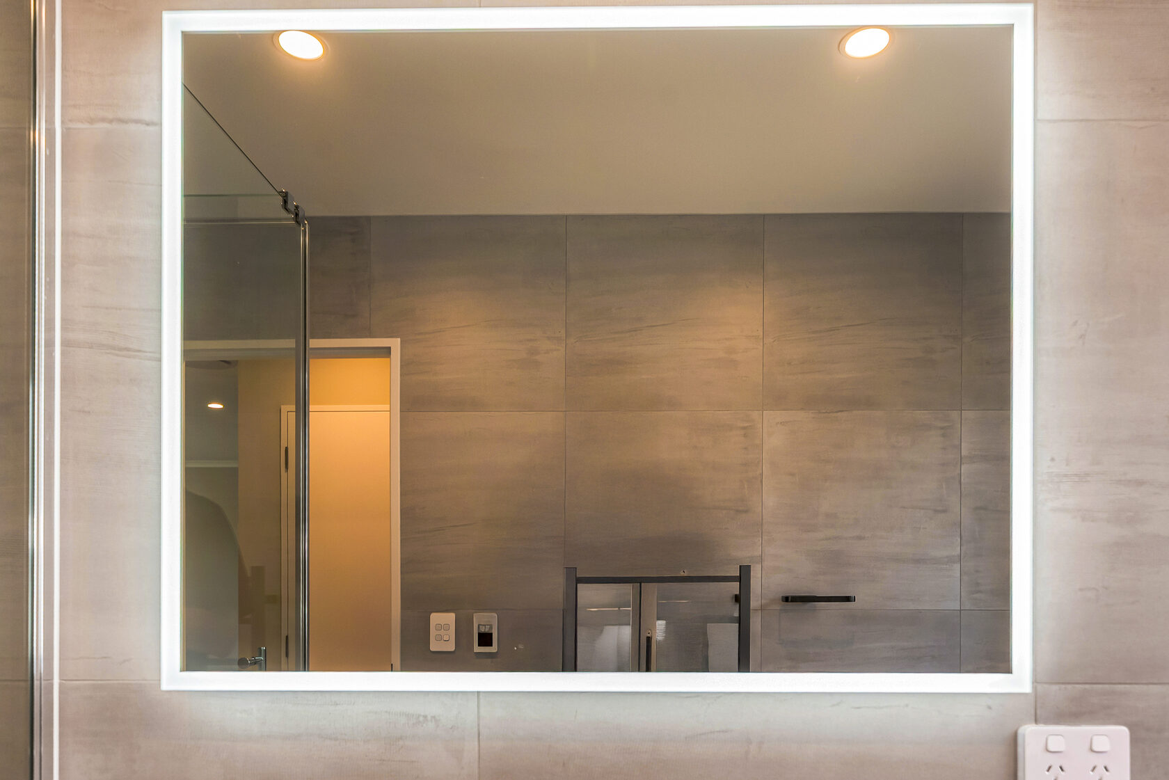 Large rectangular LED backlit bathroom mirror mounted on grey tiled wall, reflecting a glass shower enclosure and modern fixtures under recessed ceiling lights.