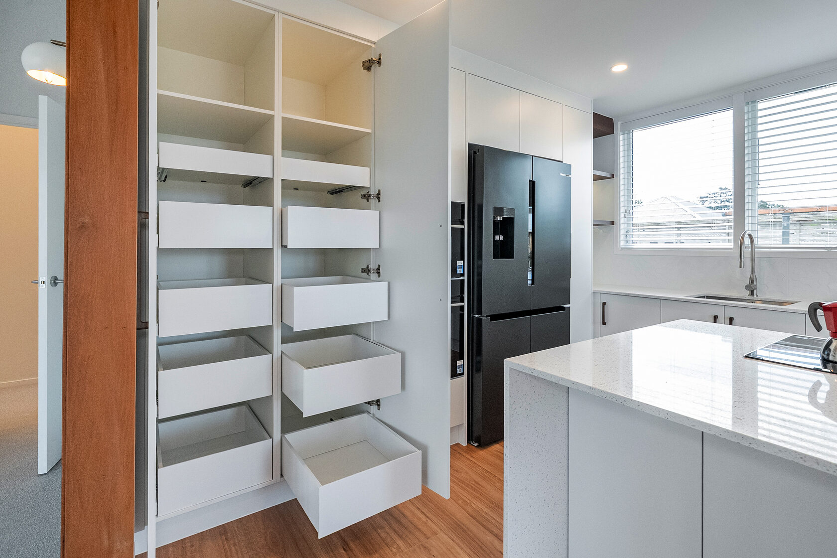 Modern kitchen with custom pull-out pantry drawers, white cabinetry, central island bench, and black double-door refrigerator.