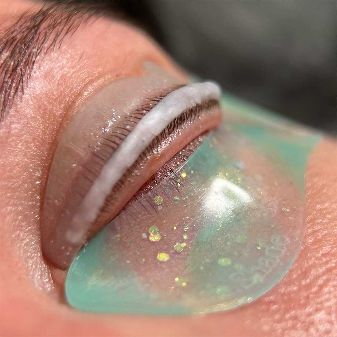 Proceso de lifting de pestañas con loción aplicada sobre el ojo, utilizando un molde y un parche verde con purpurina dorada para mejorar la fijación. Técnica especializada para conseguir un efecto natural y duradero en las pestañas.