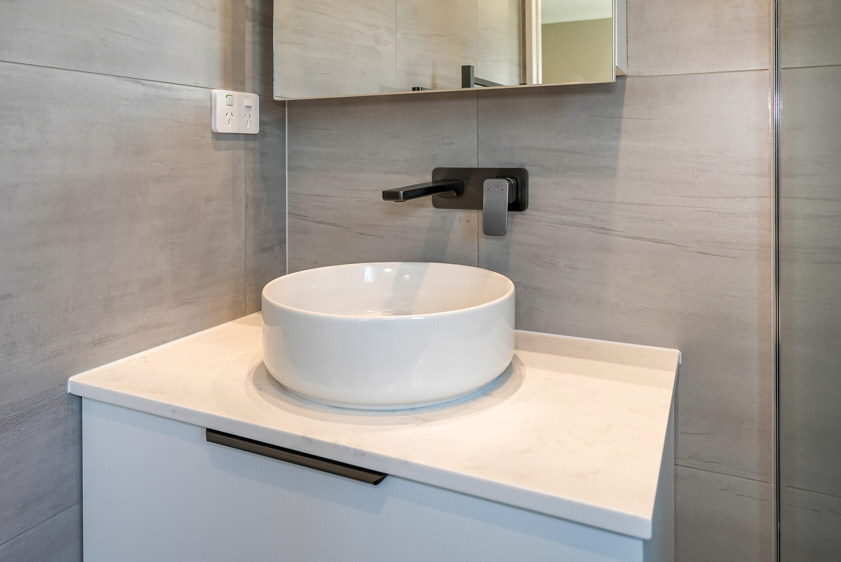 Modern bathroom vanity detail featuring a round white countertop basin, matte black wall-mounted mixer tap, light stone-look wall tiles, and a floating white cabinet with minimalist handle.