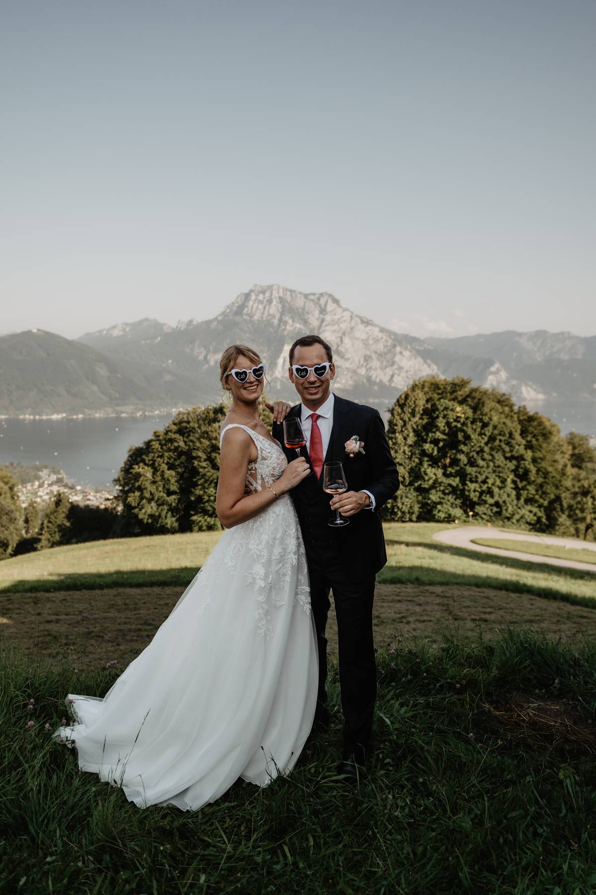 Wedding couple toasting with drinks in heart-shaped sunglasses, Austrian Alps in the background