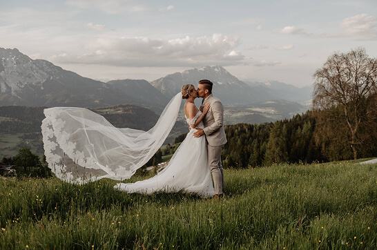 Bride and groom kissing on a hilltop with wind-blown veil — romantic mountain wedding in Schladming, Austria Alps