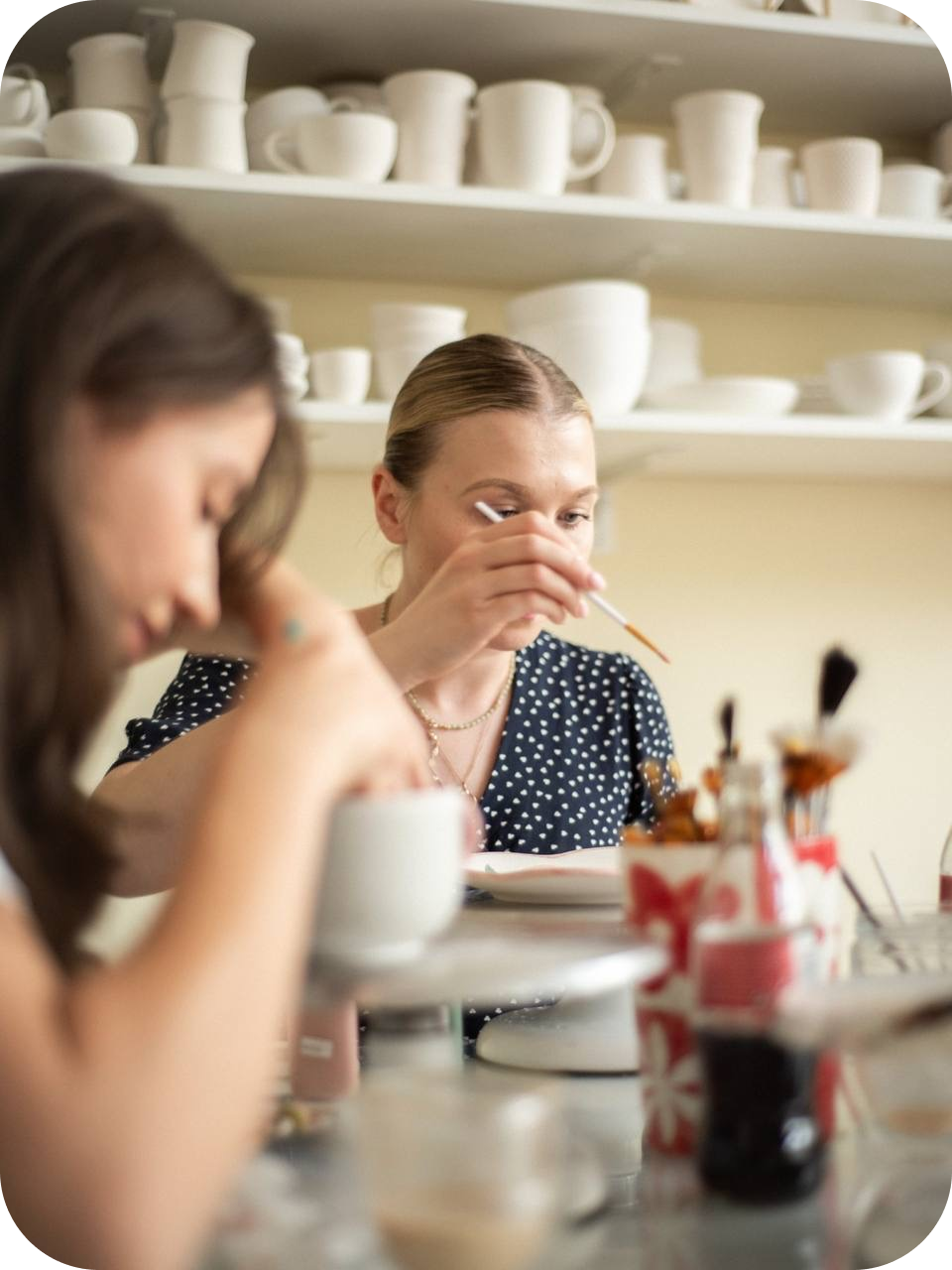 Two people painting ceramics at a table with shelves of white mugs and bowls behind them at Create Cafe in Warsaw