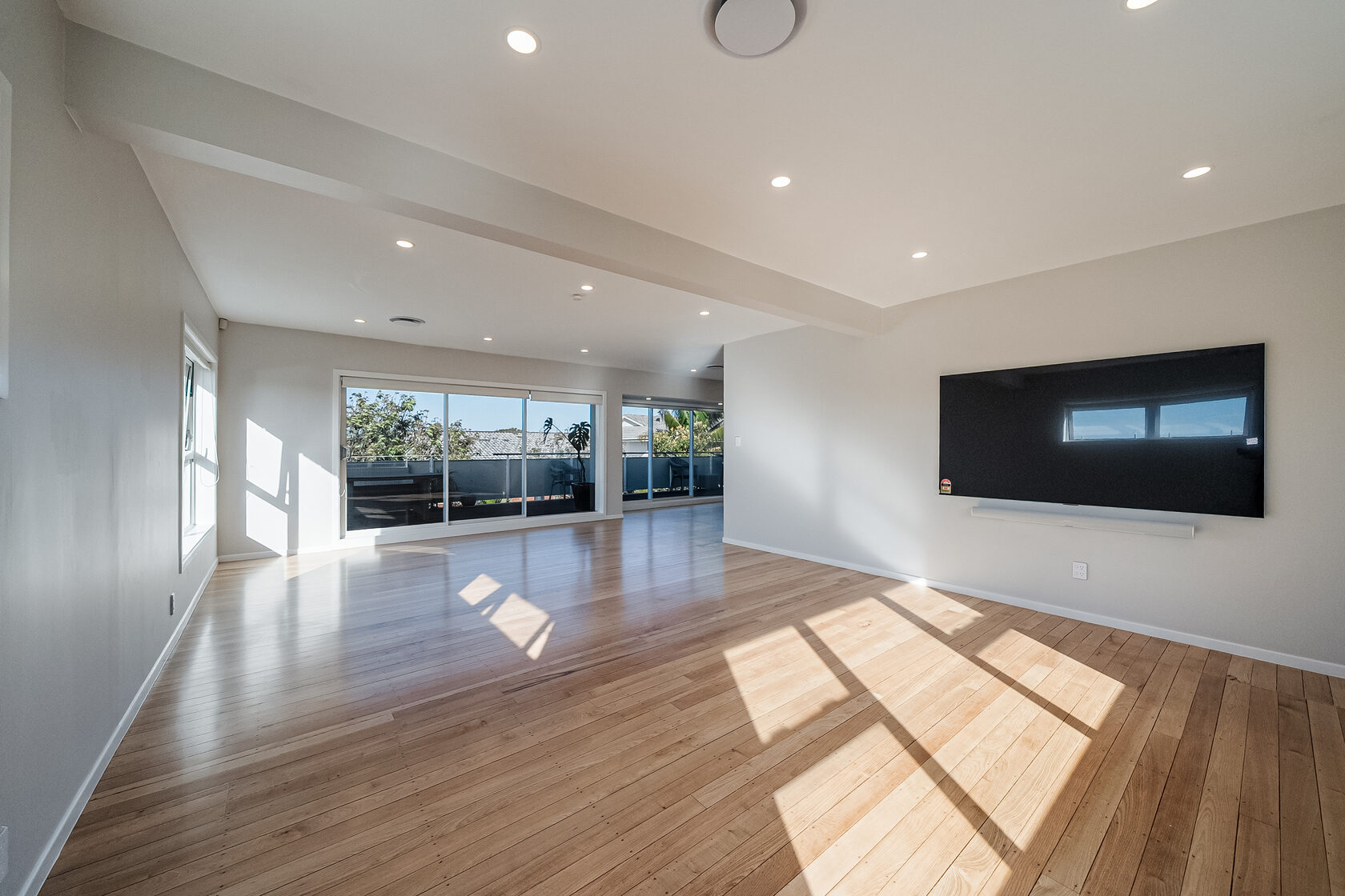Long view across the renovated Glenfield living and dining space, with continuous timber flooring and wide sliding doors to the deck