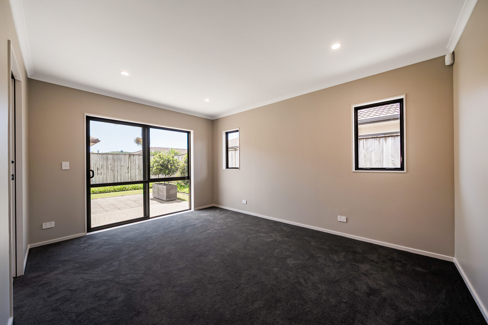 Spacious empty bedroom with dark carpet, beige walls, recessed lighting, two narrow side windows, and sliding glass doors opening to a patio and backyard.
