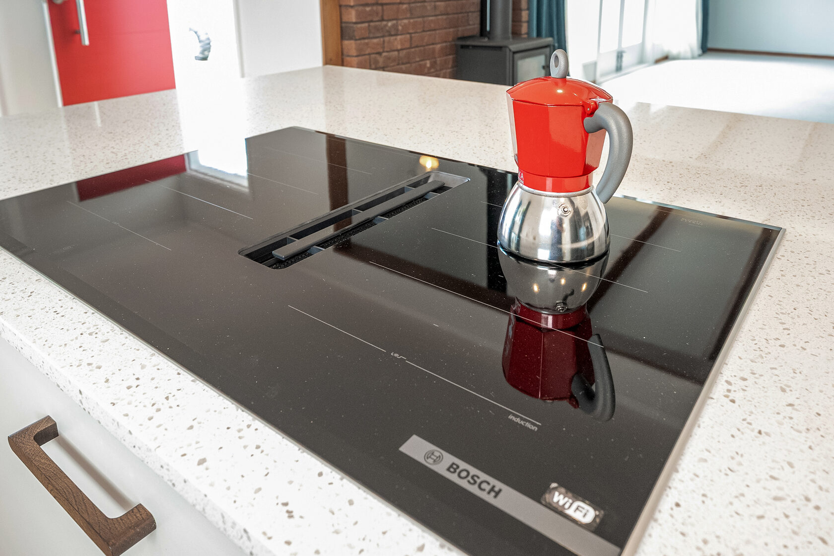 Close-up of modern induction cooktop with integrated extractor and red stovetop coffee maker on white quartz countertop.