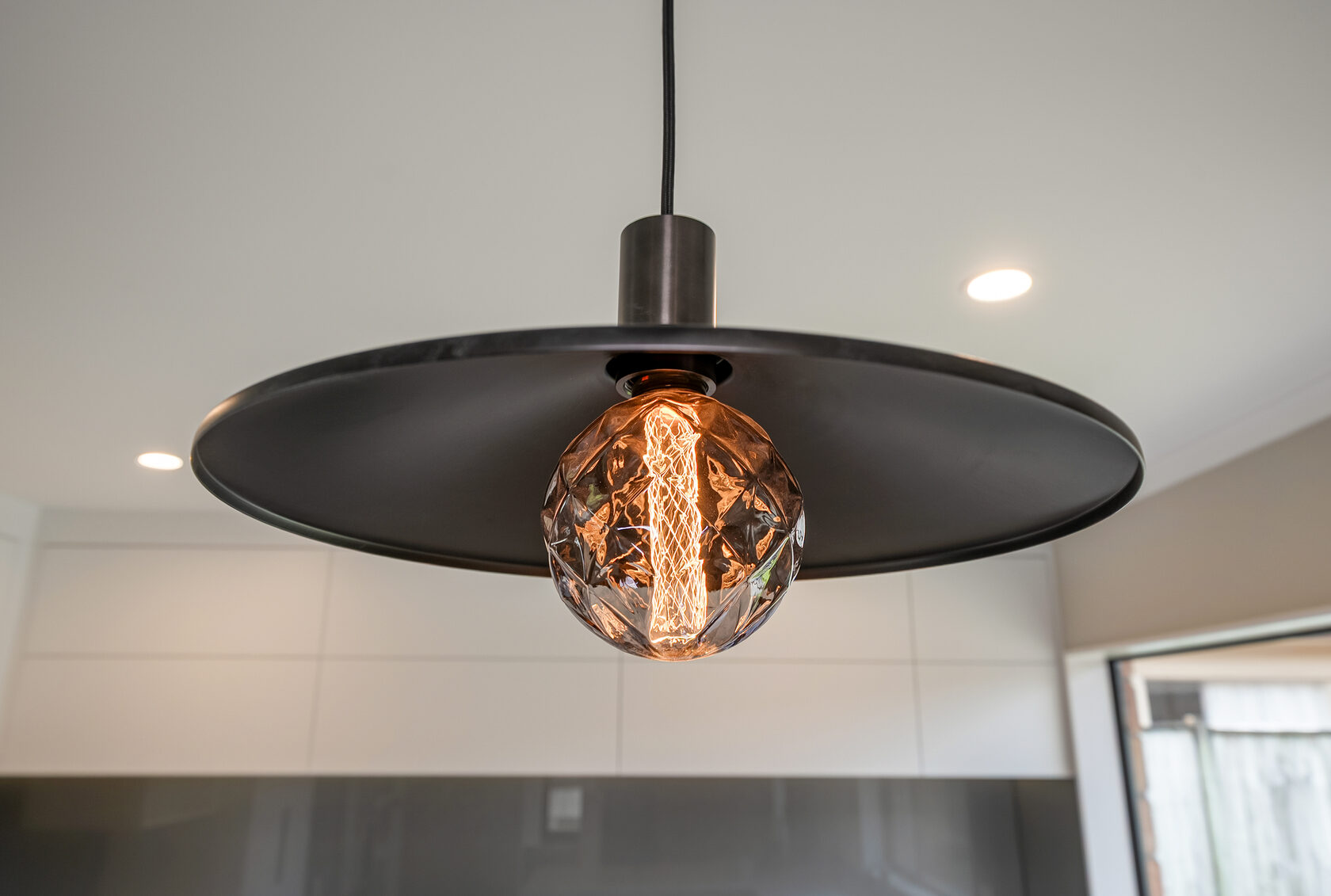 Close-up of modern black pendant light with exposed decorative filament bulb, suspended over a contemporary kitchen interior.