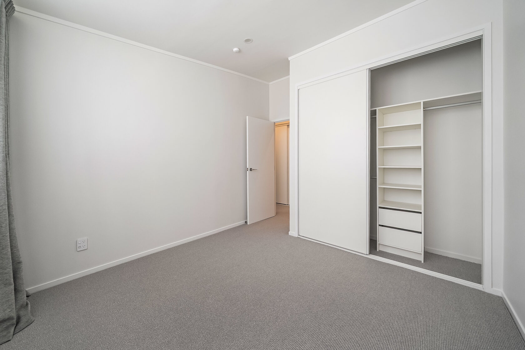 Empty bedroom with built-in wardrobe, sliding mirrored doors, carpet flooring, and neutral white walls.