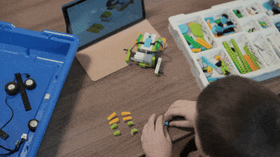 Seen from above: a child building on a wooden table, beside a tray of sorted LEGO&reg; parts, a box with wheels and a small model car