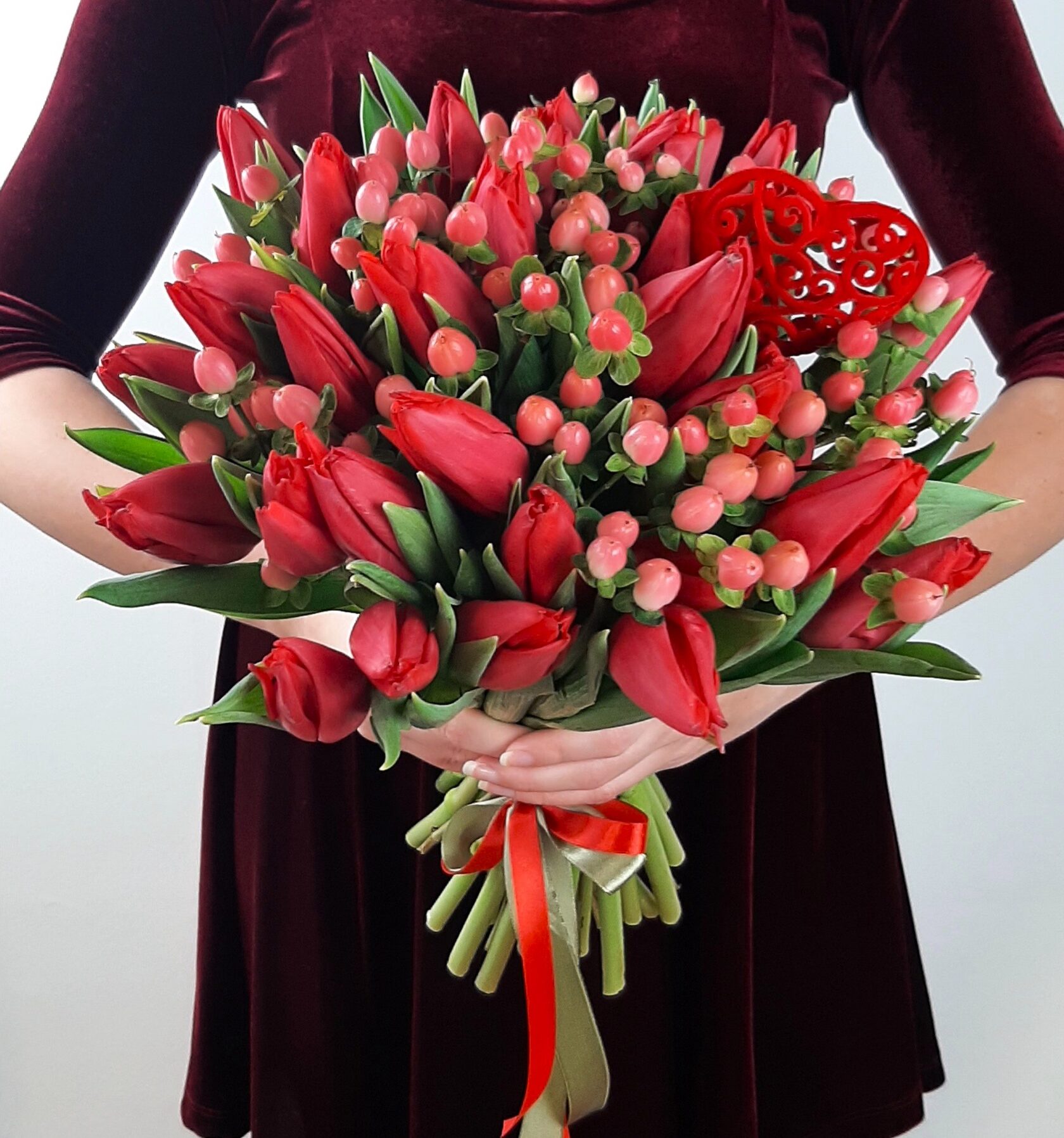 A bouquet of red tulips, accented with sprigs of pink berries (hypericum) and a decorative red heart.