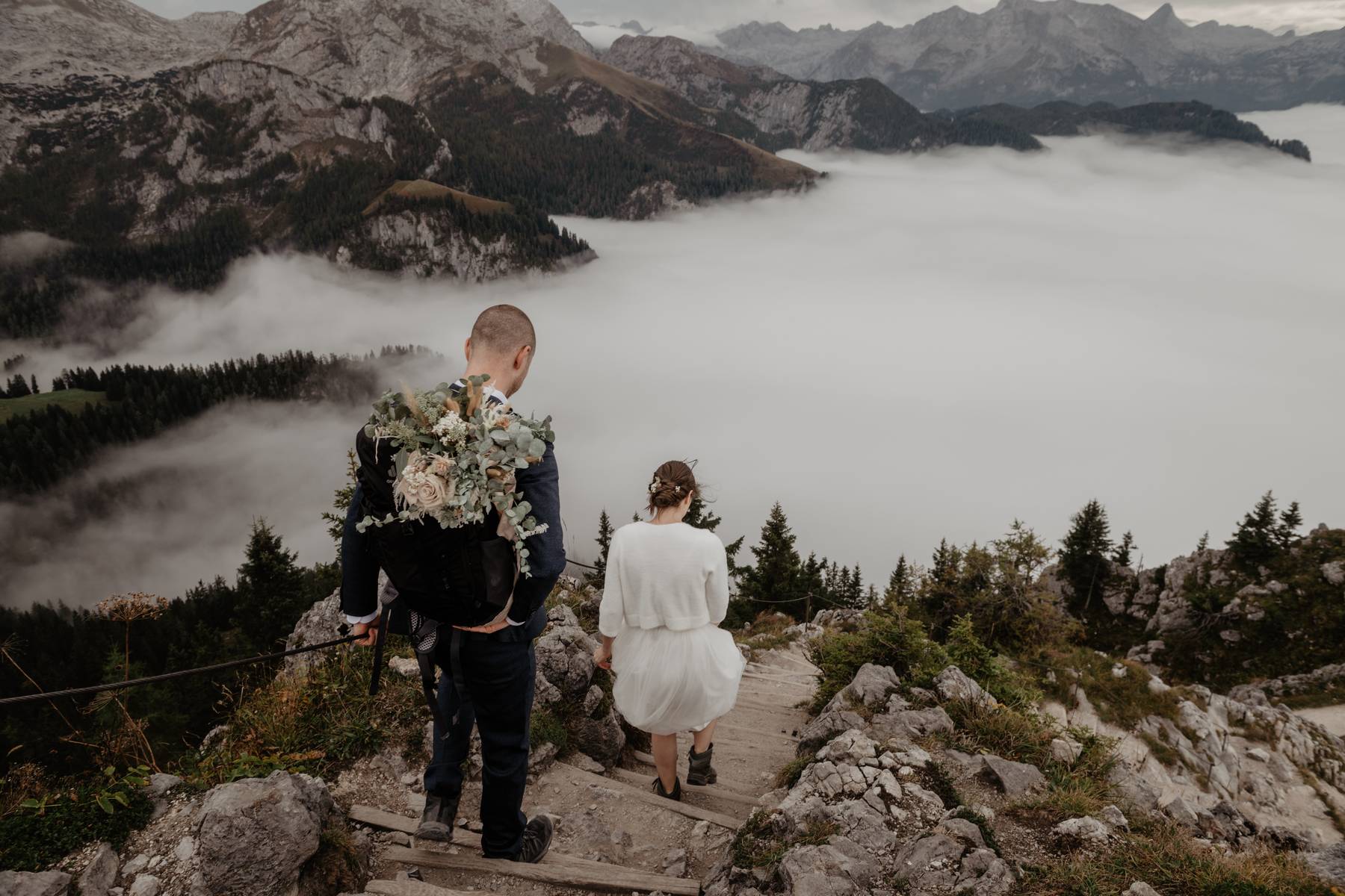 Wedding couple hiking down a rocky mountain trail above a sea of clouds, adventurous portrait