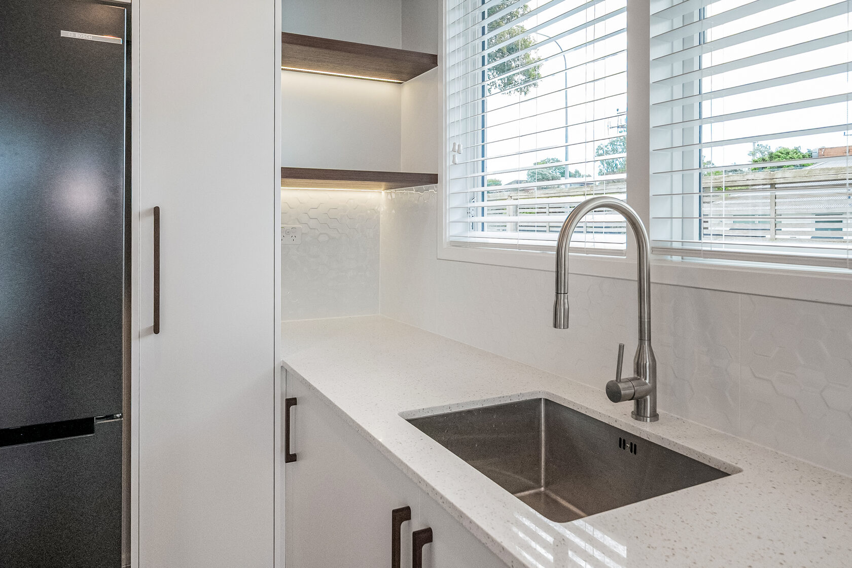 Modern kitchen sink area with stainless steel basin, brushed nickel faucet, white cabinetry, and window blinds providing natural light.