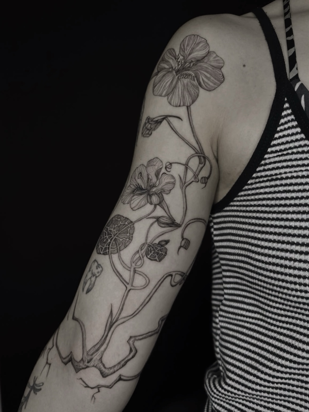 Upper-arm tattoo of two open flowers, buds, round veined leaves and winding stems with angular branches.