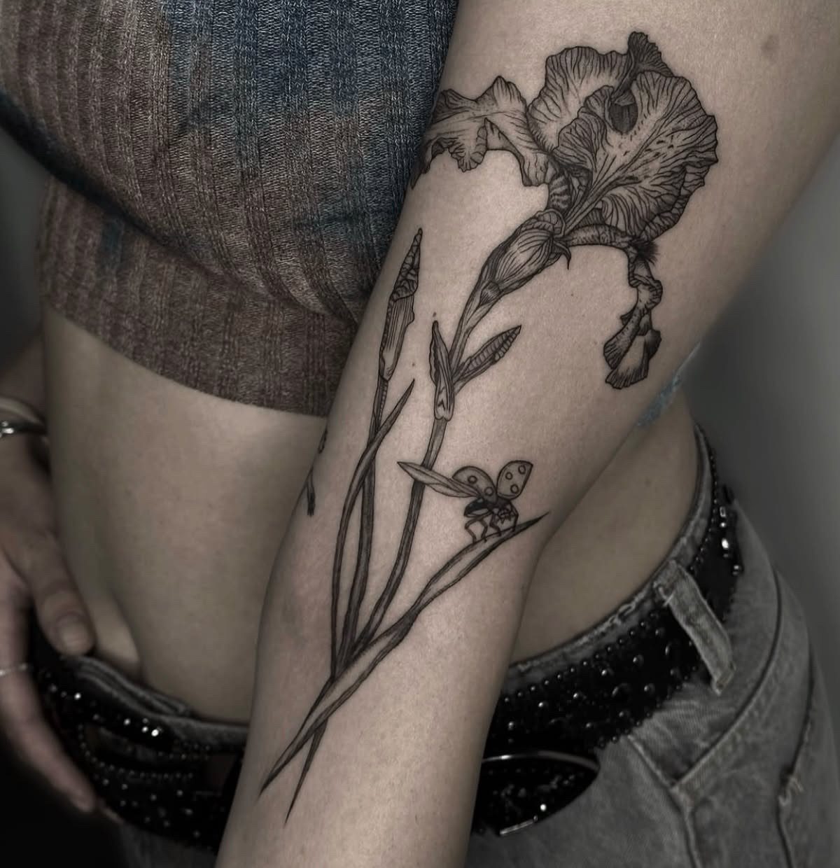 Arm tattoo of a large flower with ruffled petals, a bud, narrow leaves and a small spotted beetle on one leaf.
