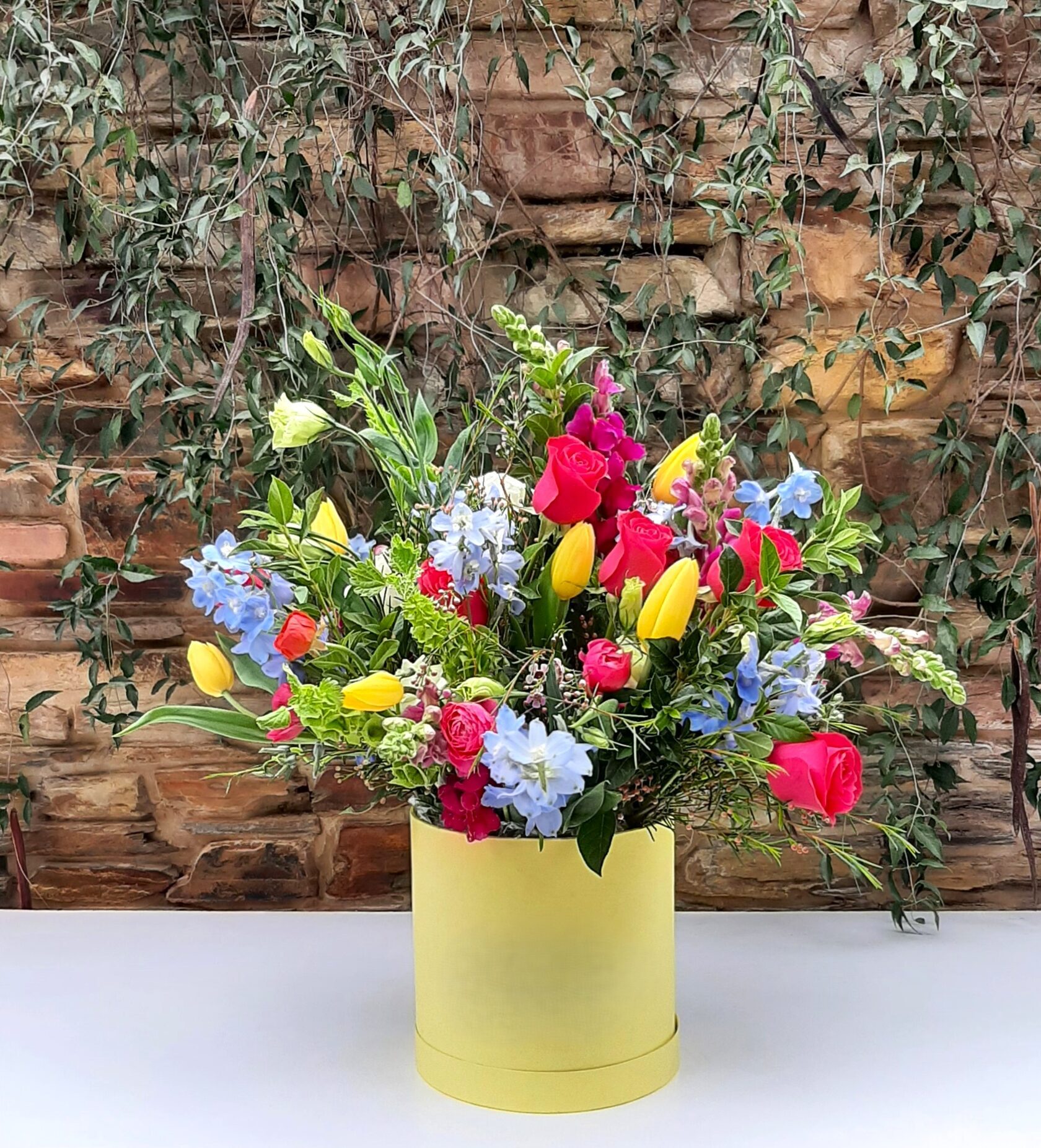 An arrangement in a yellow hat box, consisting of pink roses, yellow tulips, and blue delphiniums.