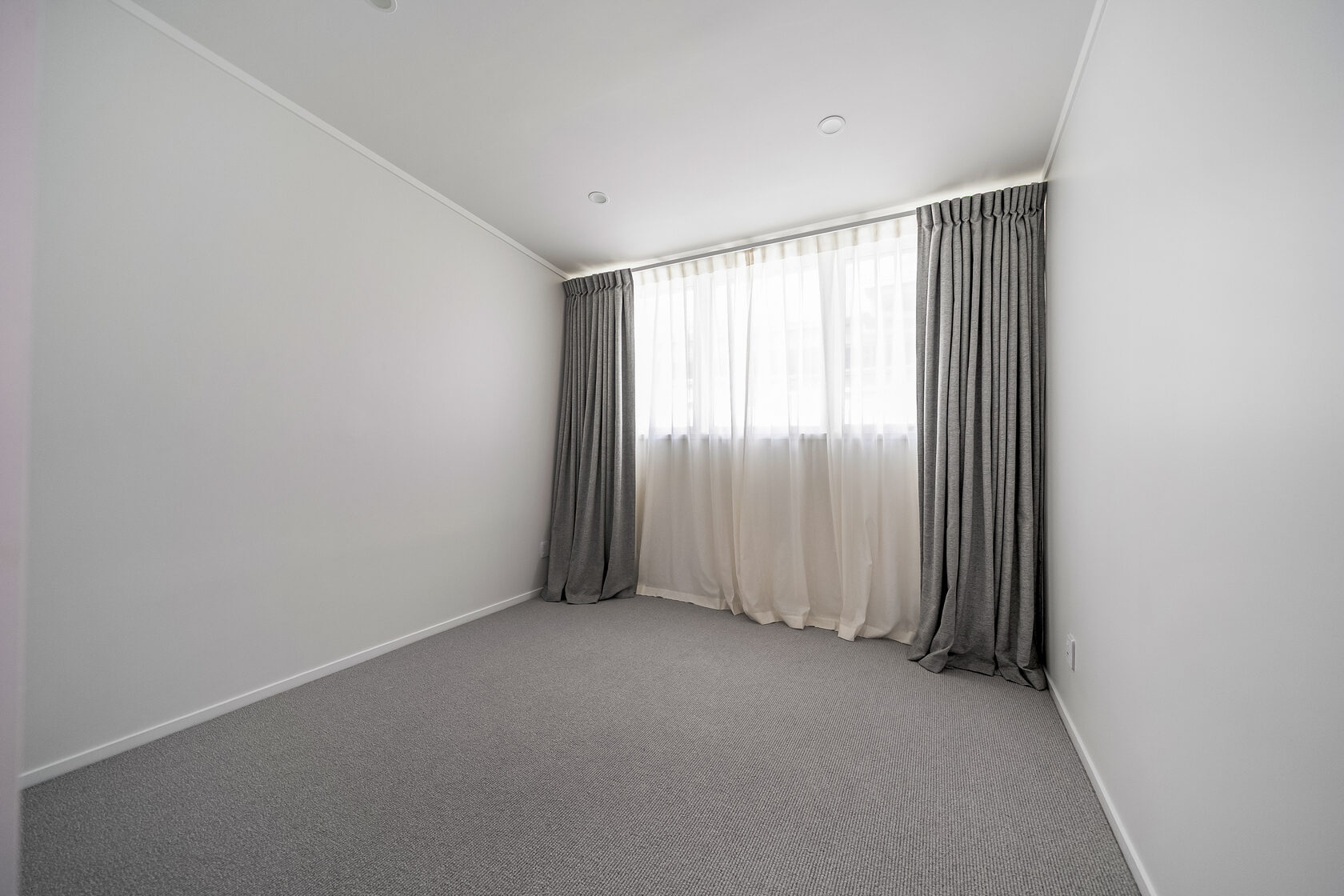Empty bedroom with neutral walls, carpet flooring, recessed ceiling lights, and large window with sheer and blackout curtains.