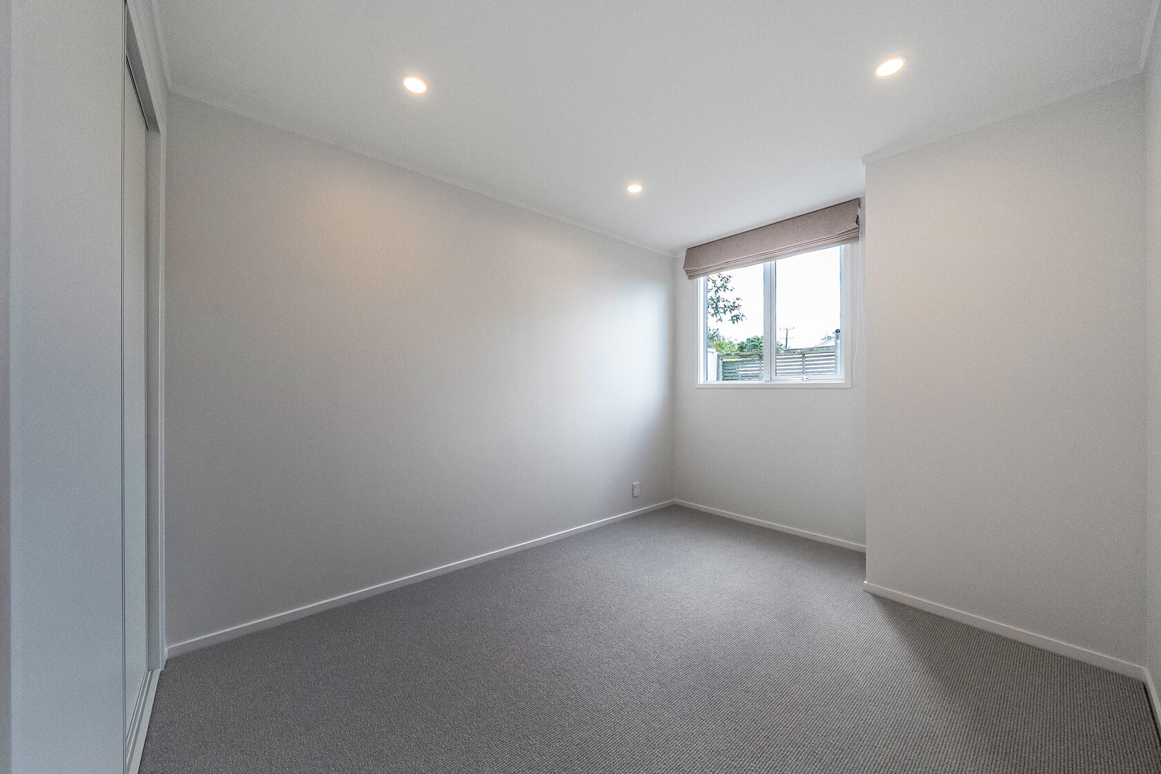 Small bedroom with neutral walls, carpet flooring, recessed ceiling lights, and window with roller blind.