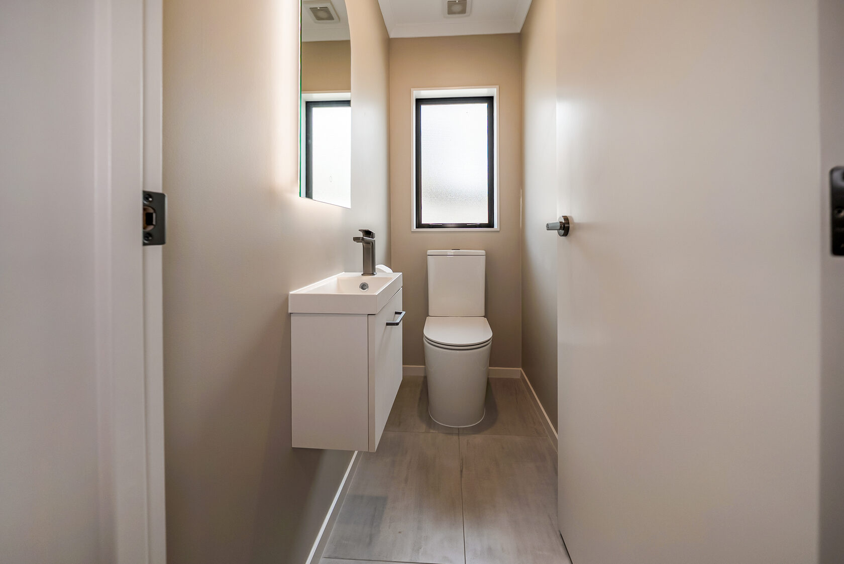 Compact guest toilet with neutral beige walls, slim floating vanity and mixer tap, modern white toilet suite, frosted window for privacy, and light grey floor tiles.