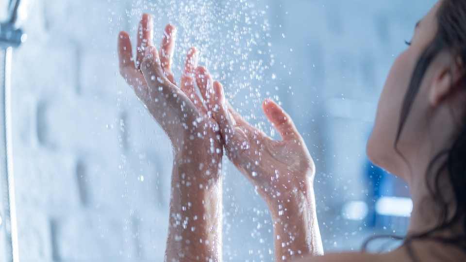 The image shows a woman testing water hardness in the shower by observing water flow and lather formation, a simple method to detect hard water at home