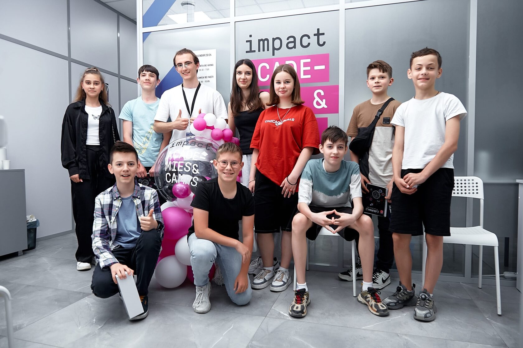 Een groep van zo&rsquo;n twaalf leerlingen en hun docent op de foto voor de muur van Impact Academies & Camps, naast roze en witte ballonnen