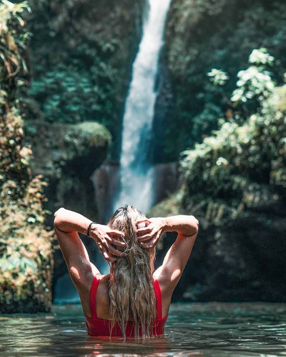 Mujer en traje de baño rojo descansando en una poza natural junto a una esbelta catarata