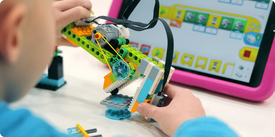 A child&rsquo;s hands holding a green and orange LEGO&reg; model with gears and a jaw, in front of a tablet in a pink case