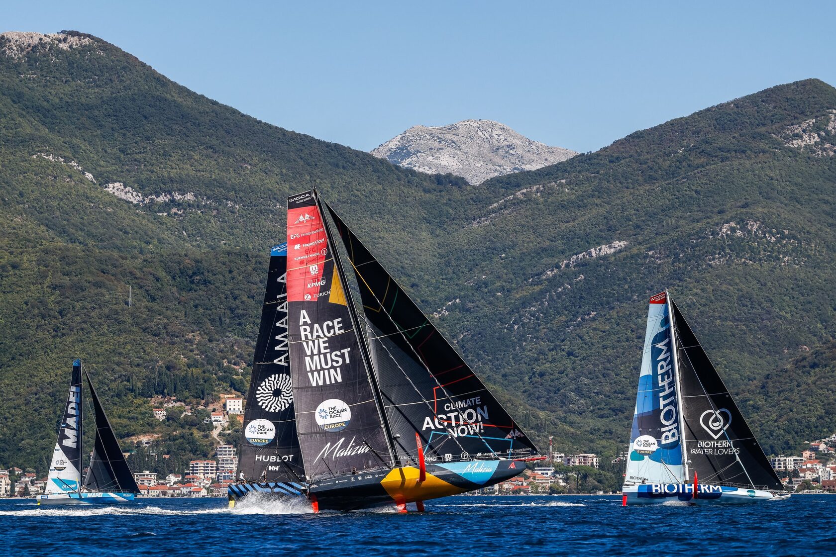 IMOCA yachts racing against a mountainous coastal backdrop during The Ocean Race Europe