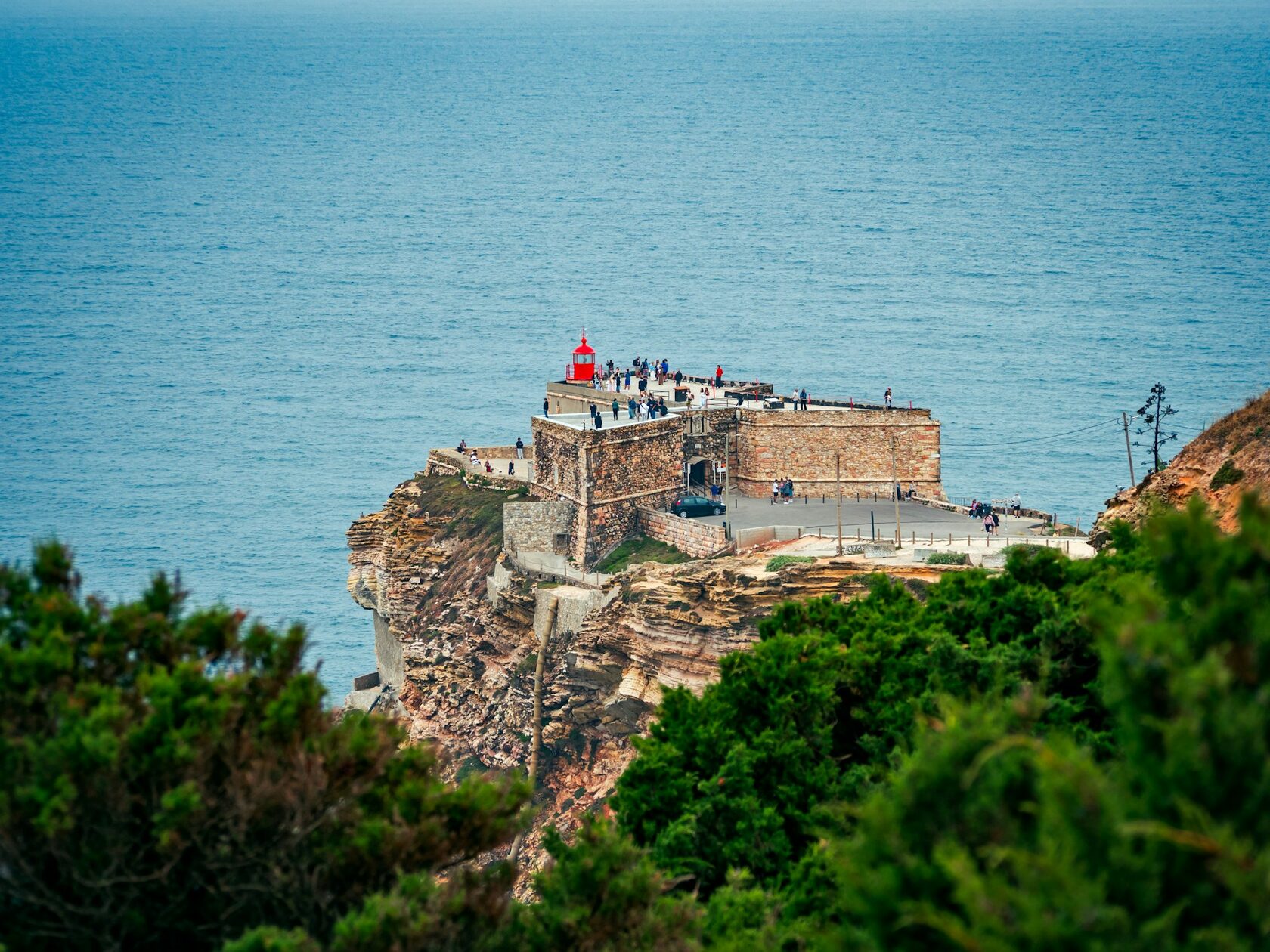 The red lighthouse fort at Nazaré on its cliff, a giant Atlantic wave breaking behind it