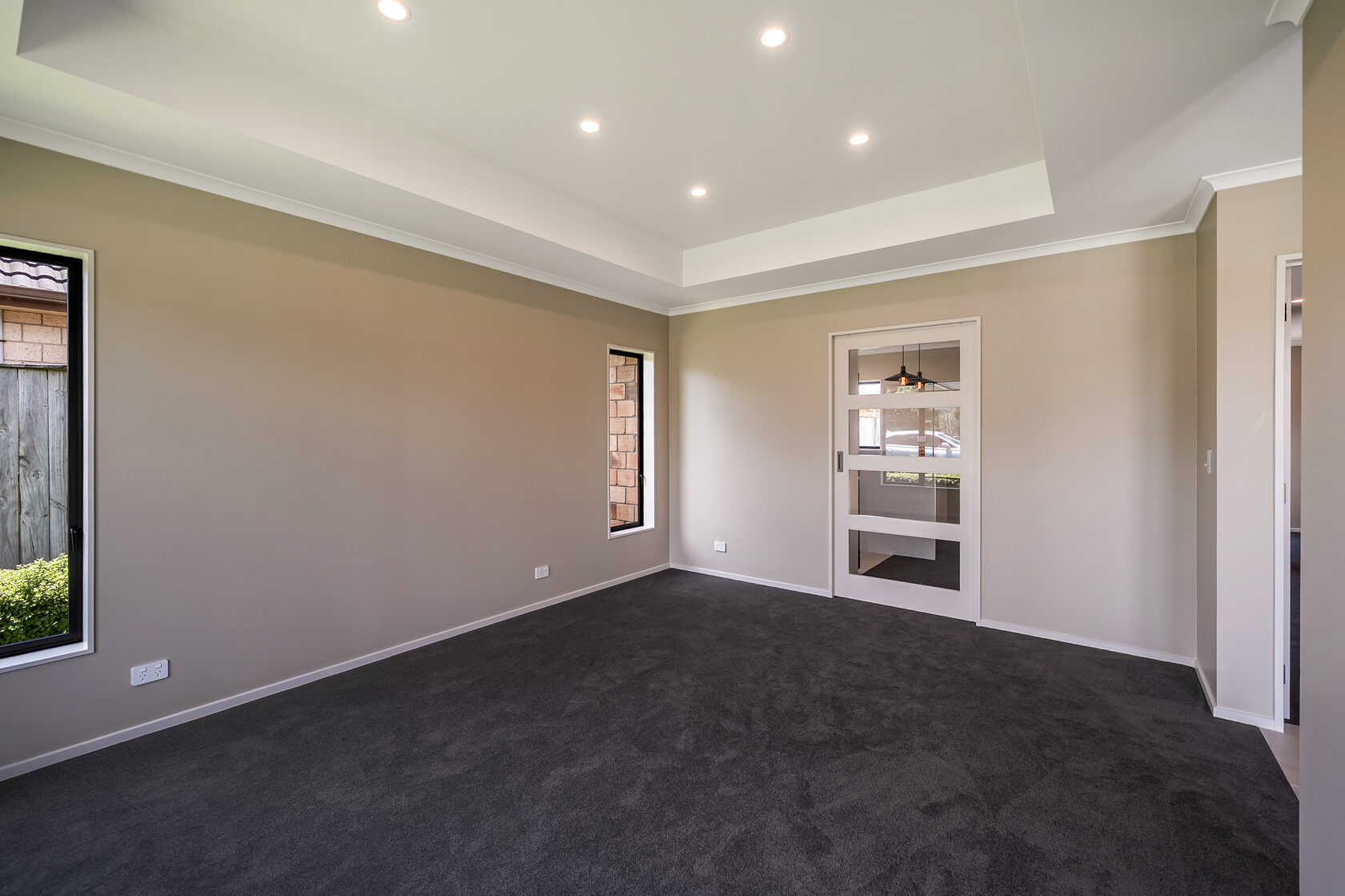 Empty carpeted room with recessed ceiling and downlights, neutral walls, side window, and glass-panel door leading to the kitchen area.