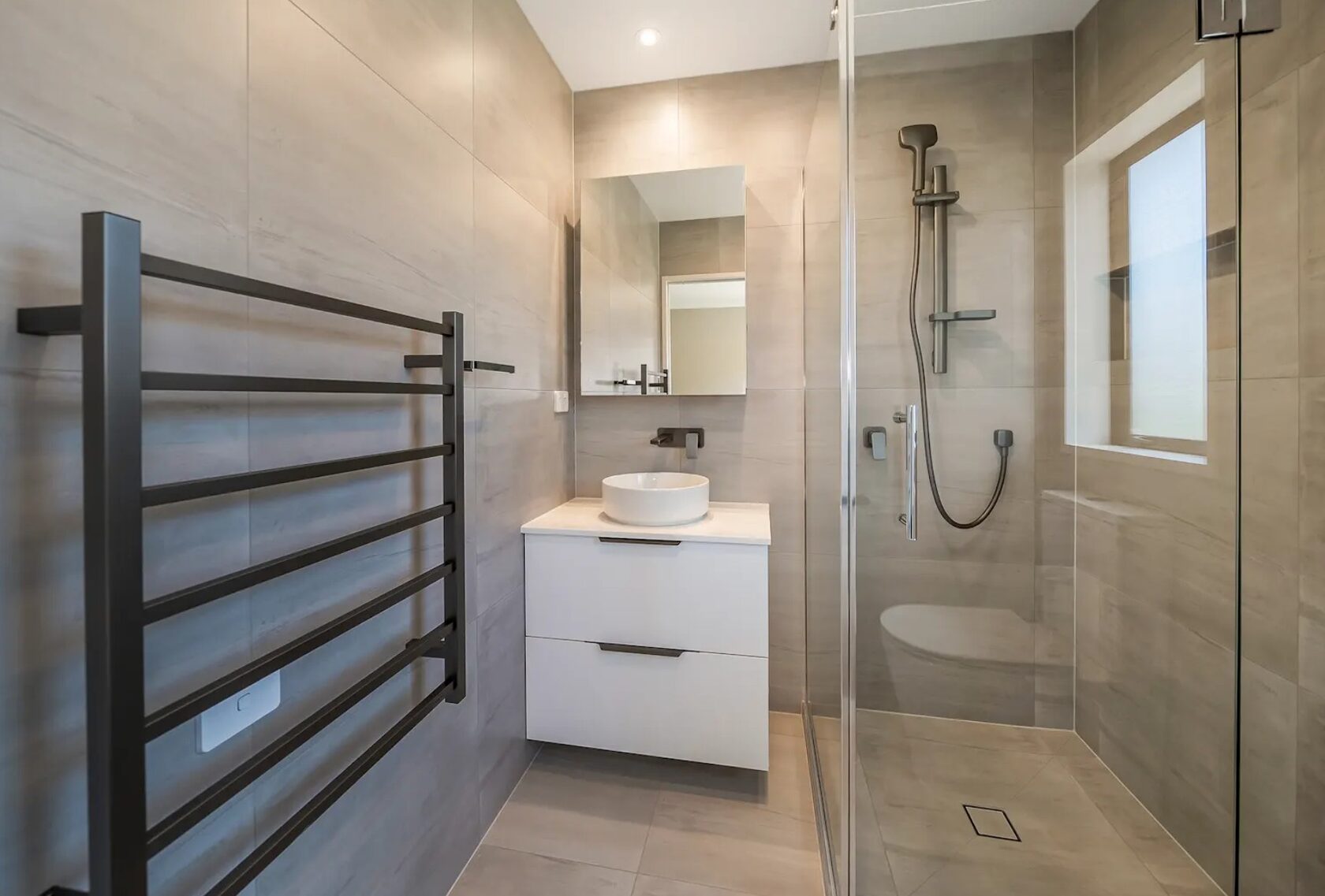 After: Renovated ensuite with grey wall tiles, glass shower, white vanity and black heated towel rail