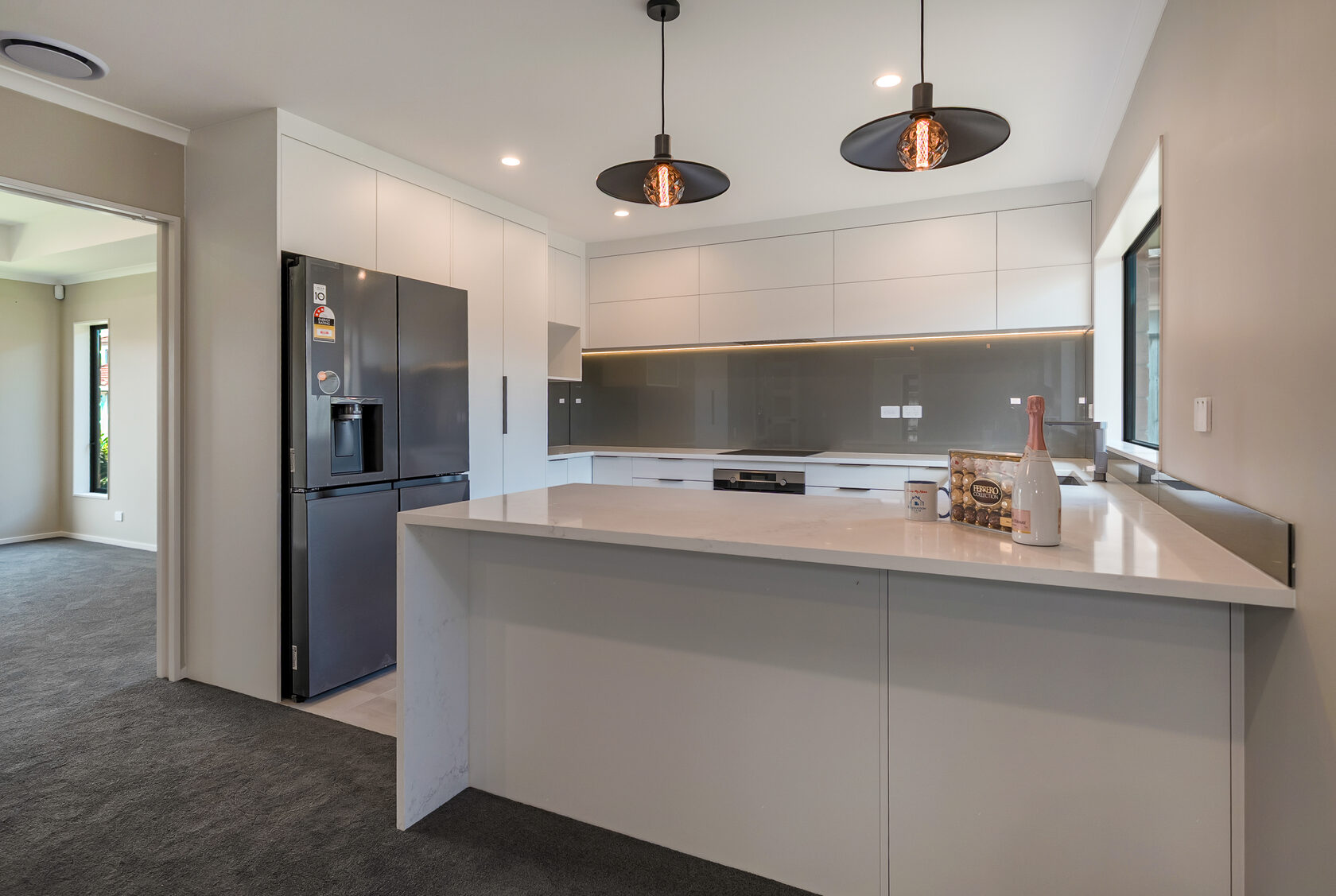Modern white kitchen with peninsula bench, grey glass splashback, black double-door fridge, and two contemporary pendant lights with exposed filament bulbs.