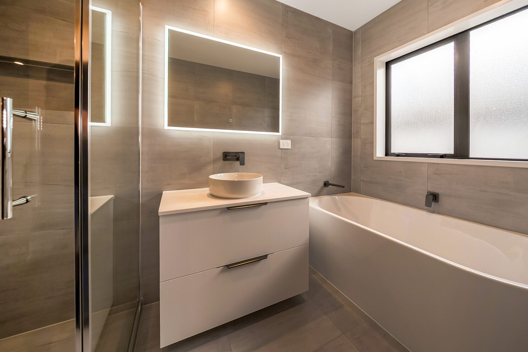 Modern bathroom with floating white vanity, round vessel basin, matte black wall-mounted tapware, LED backlit mirror, and a sleek freestanding bathtub set against large-format grey tiles.